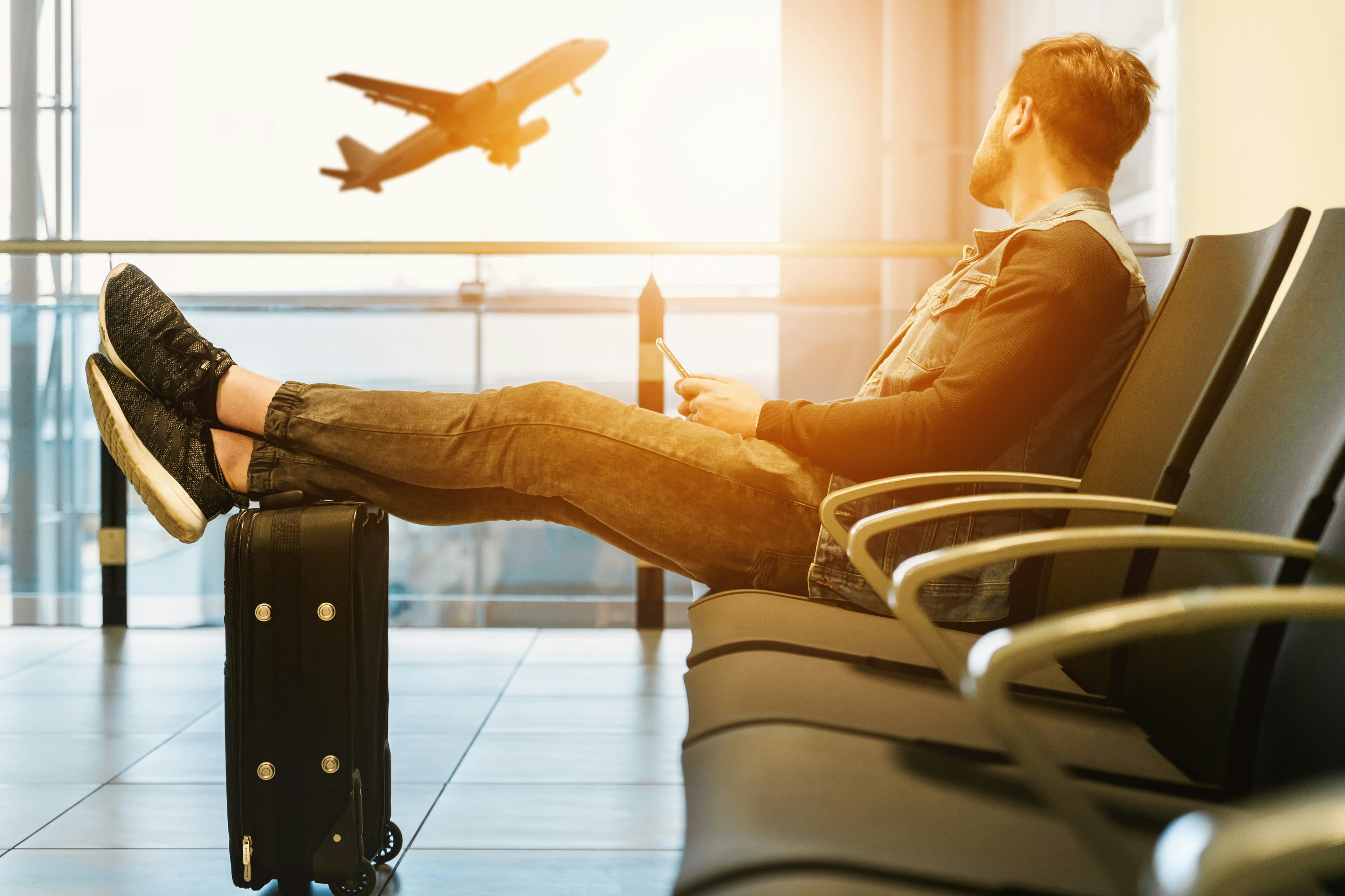 A traveler with his feet up on a suitcase, watching a plane take off through an airport window