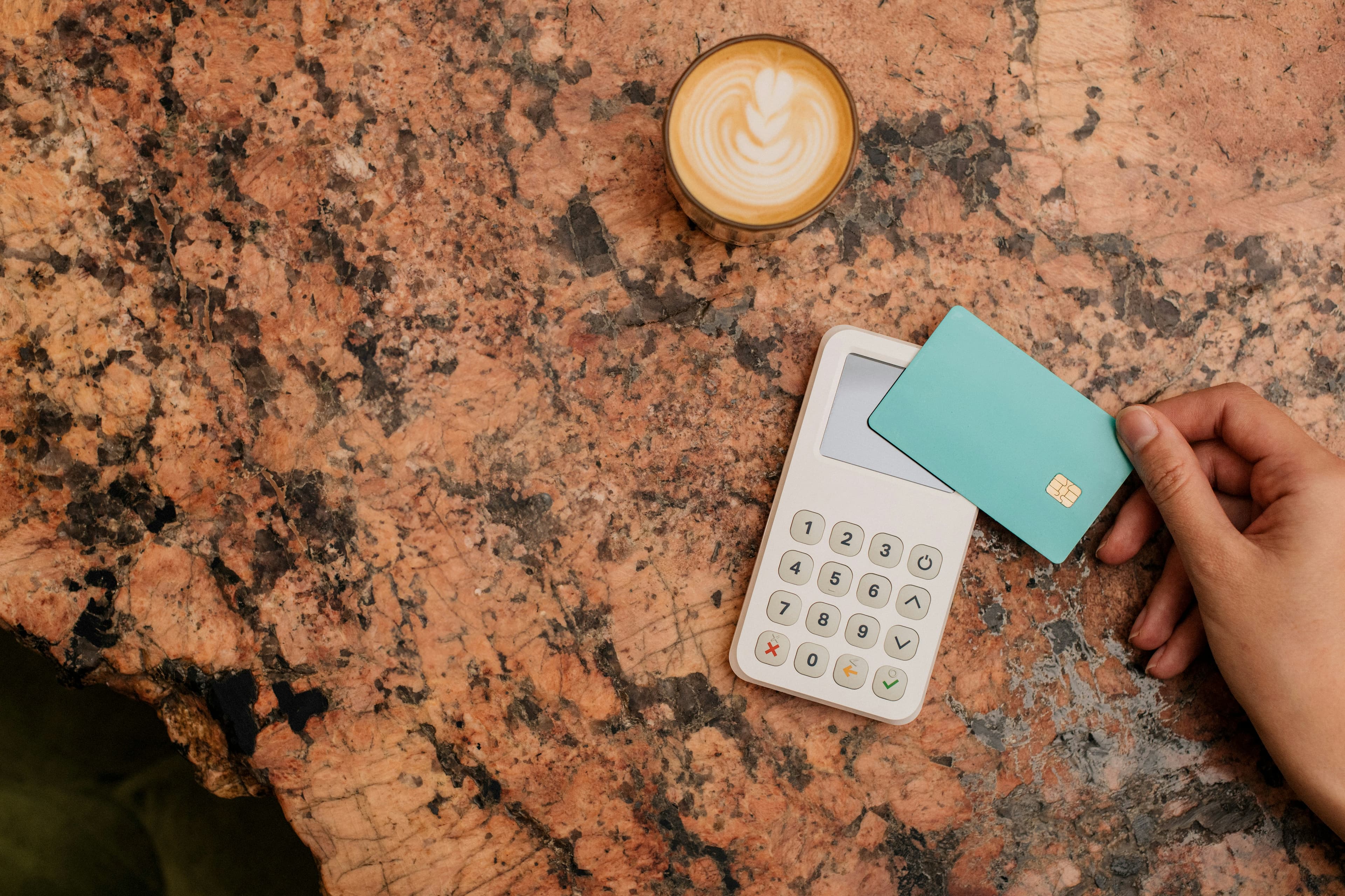 A hand holding a bank card beside a payment terminal and a coffee on a stone table