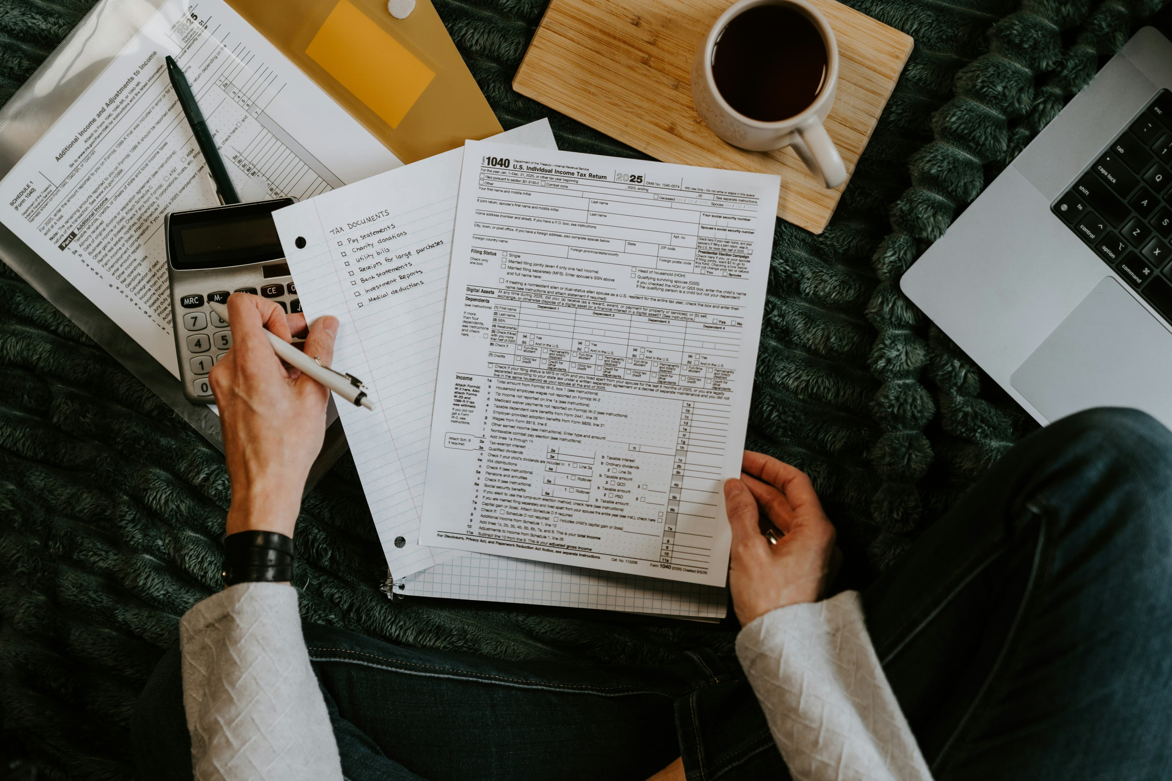 A person filling in tax forms beside a calculator, a laptop and a cup of coffee