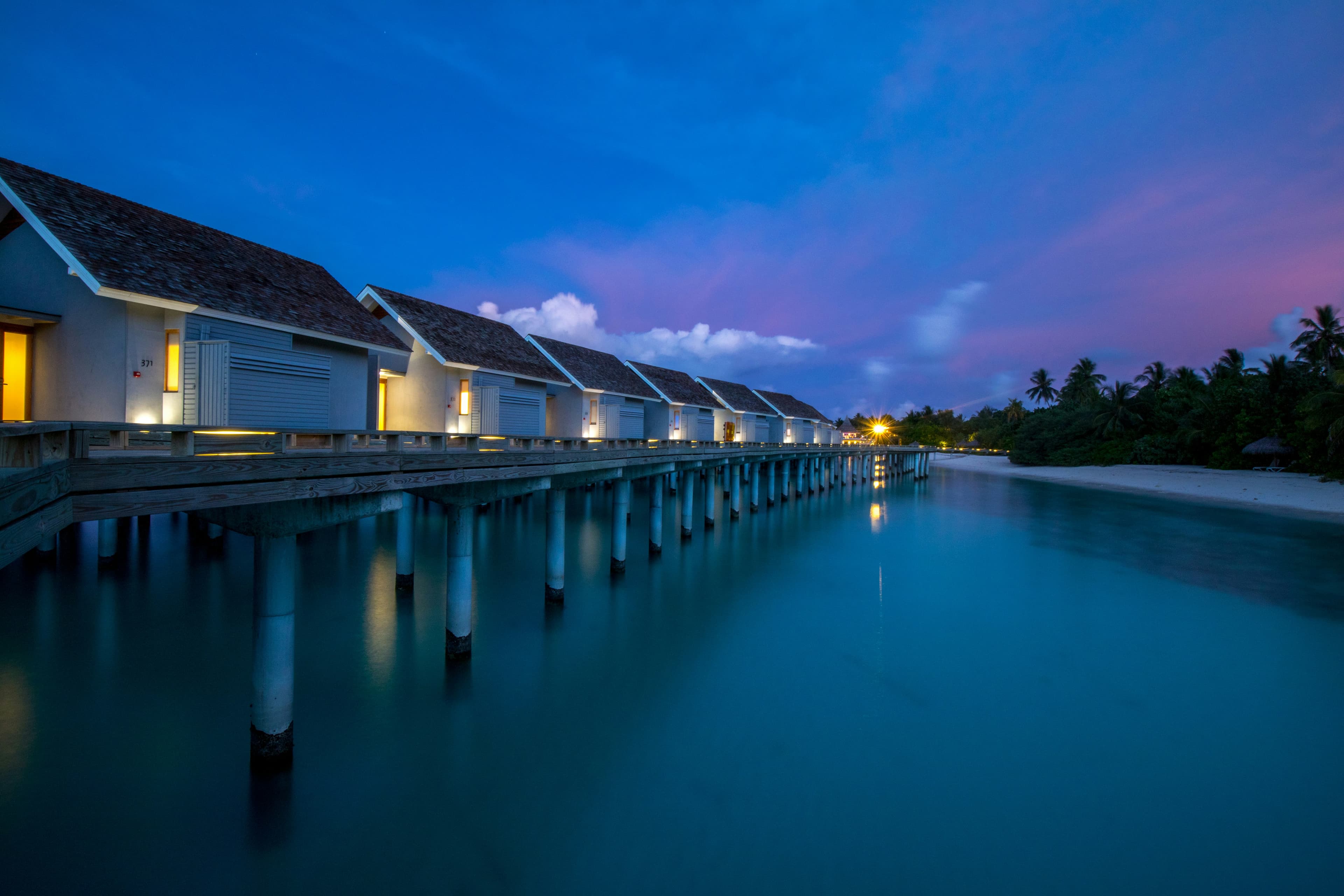 Overwater villas lit at dusk along a jetty, under a deep blue evening sky